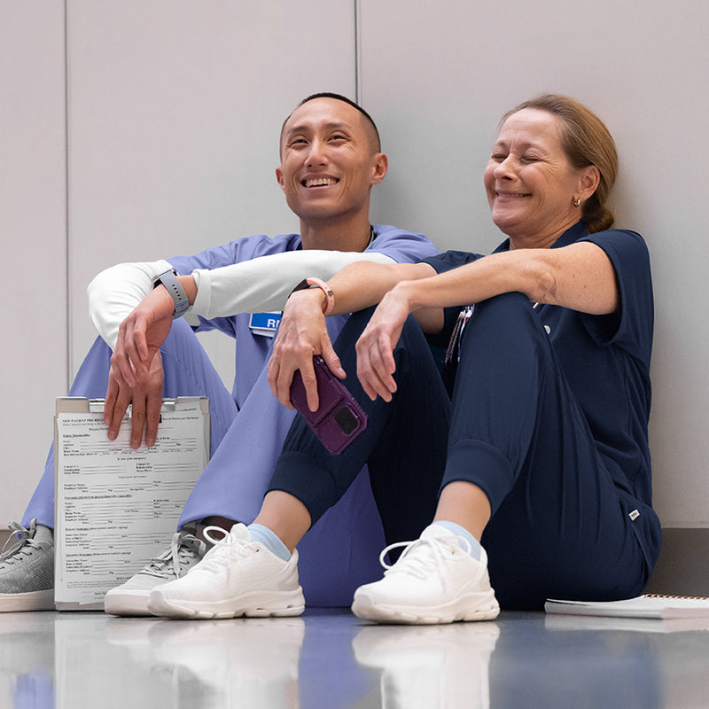 Two nurses in scrubs sitting on a floor with a document in front of them, wearing matching outfits.