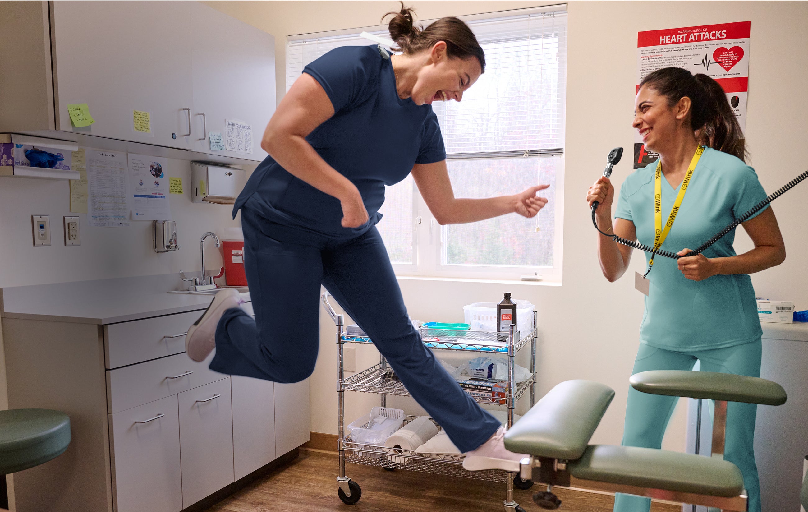 Two healthcare professionals in a medical office setting in scrubs, one jumping and the other holding an instrument