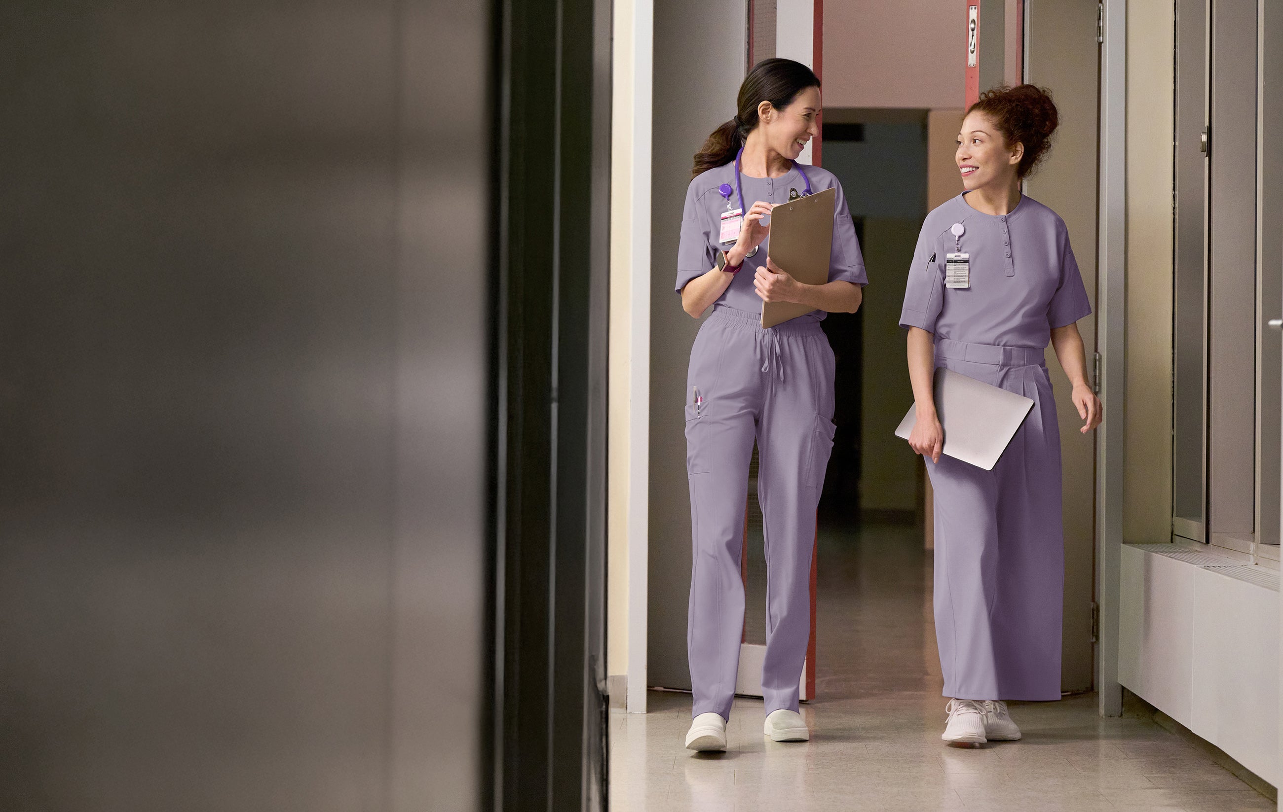 Two nurses in purple scrubs walking down a hospital hallway.
