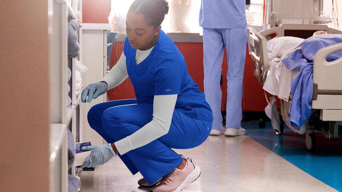 Person in royal blue scrubs crouching in a hospital room with medical equipment.