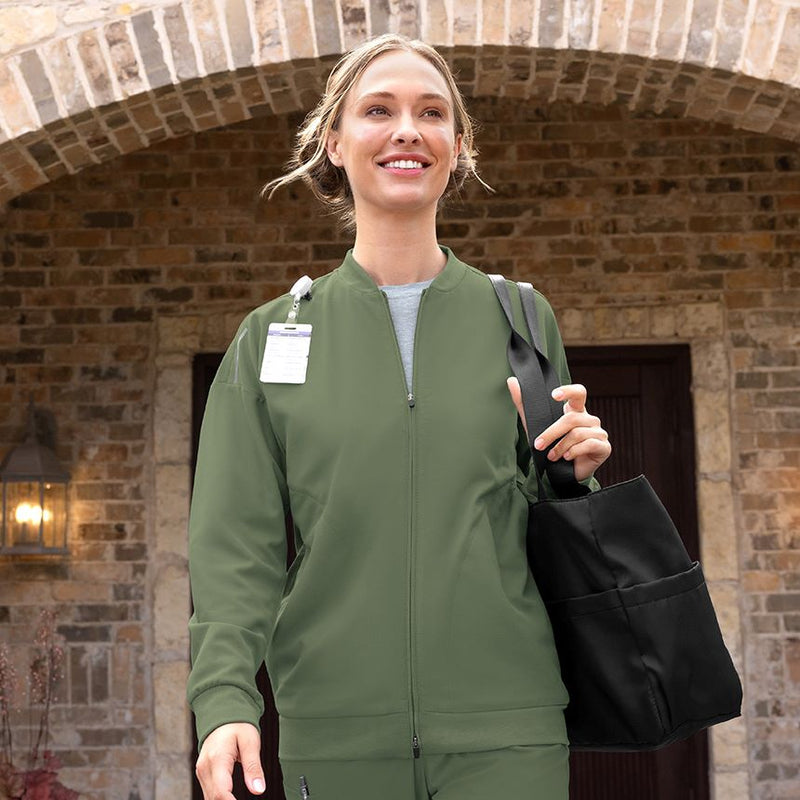 Woman in green medical jacket holding a black bag against a brick wall.