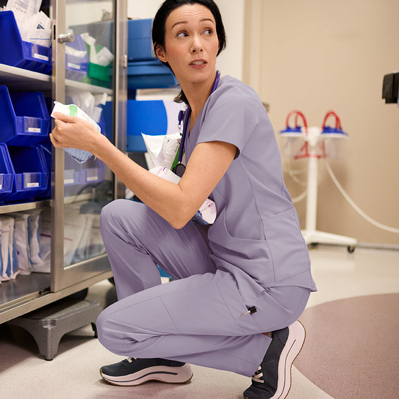 Nurse in purple scrubs kneeling in a medical setting with shelves and equipment in the background