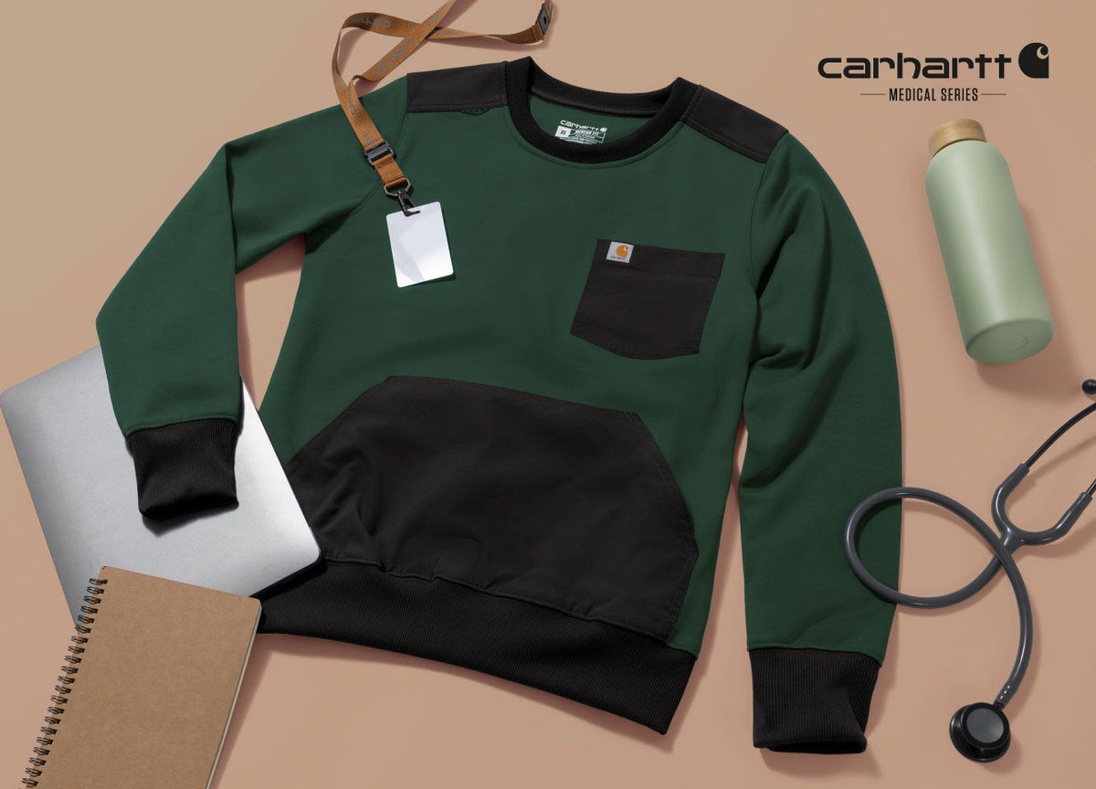 A Carhartt Medical Series crewneck sweatshirt in forest green and black, styled in a flat lay with a stethoscope, notebook, laptop, water bottle, and lanyard on a beige background.