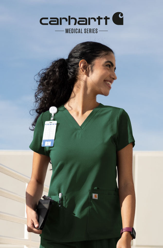 A woman wearing dark green medical scrubs stands outdoors under a blue sky, with the Carhartt Medical Series logo at the top of the image. She has a badge clipped to her collar and a pen in the pocket of the scrub top.