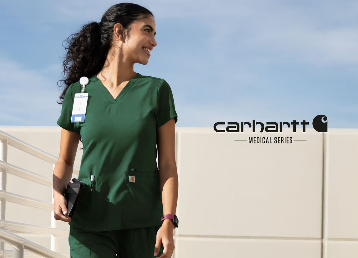 A woman wearing dark green medical scrubs walks outdoors against a light blue sky and beige wall, with the Carhartt Medical Series logo printed on the right side of the image.