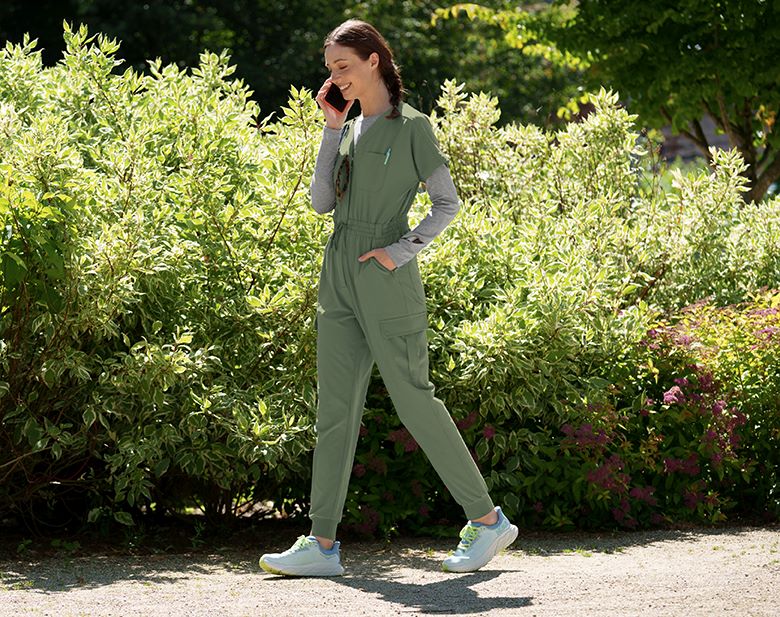 A woman wearing a jogger scrub Jumpsuit walks next to bushes while talking on the phone.