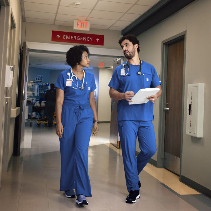 Two nurses in blue carhartt scrubs walking down a hospital hallway.
