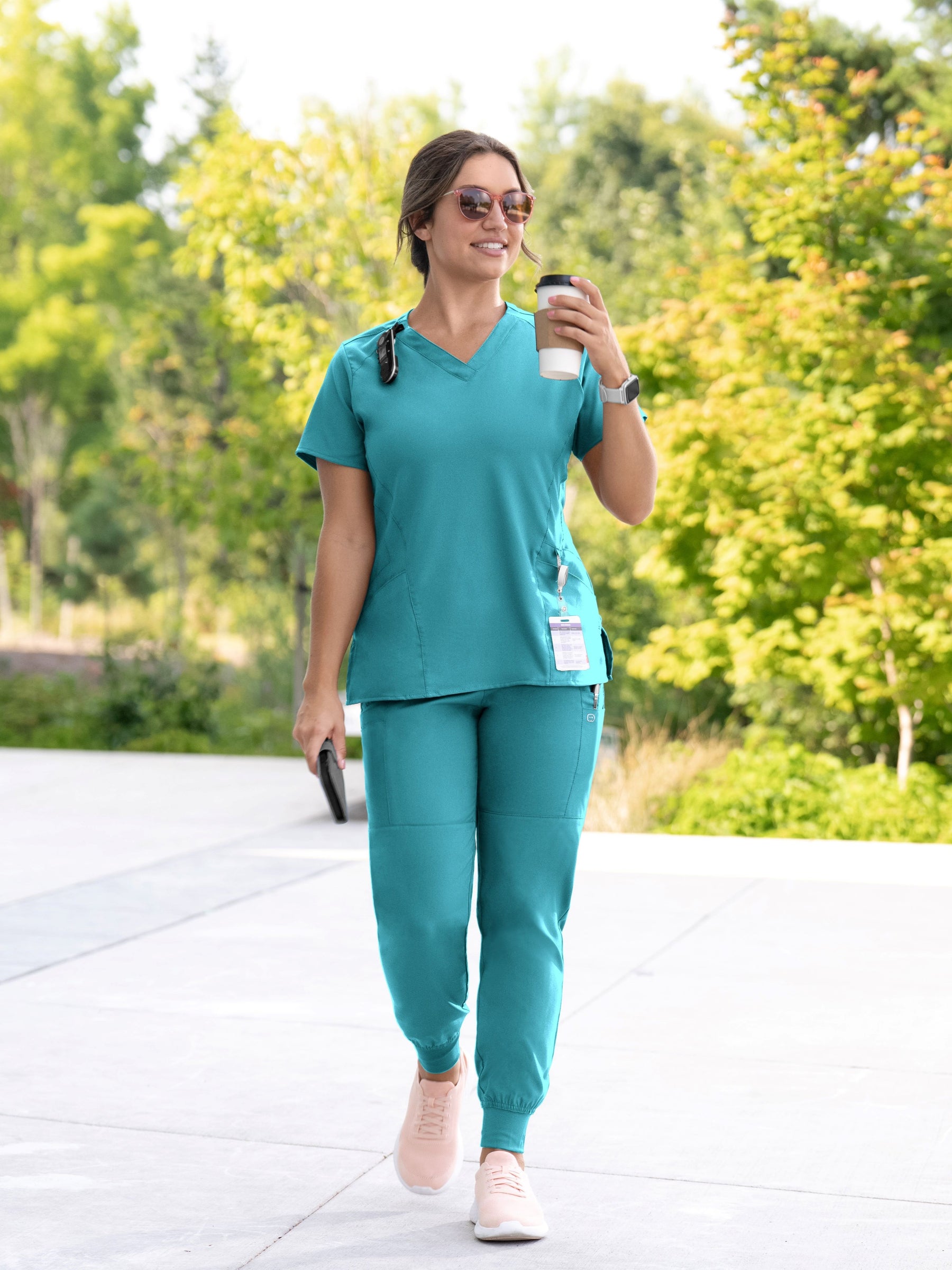 Woman in teal scrubs walking outdoors holding a coffee cup