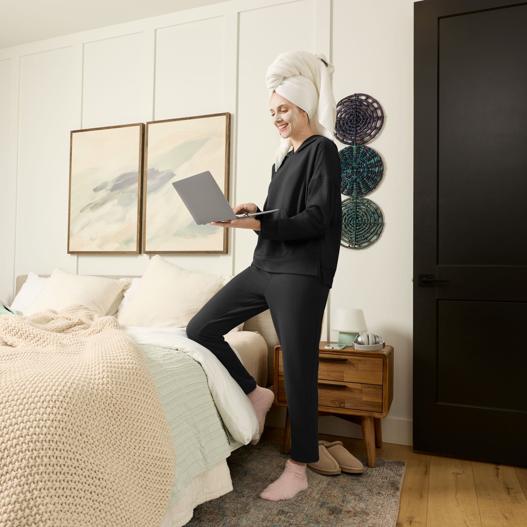 Person in a bedroom using a laptop wearing black loungewear set with a towel on their head